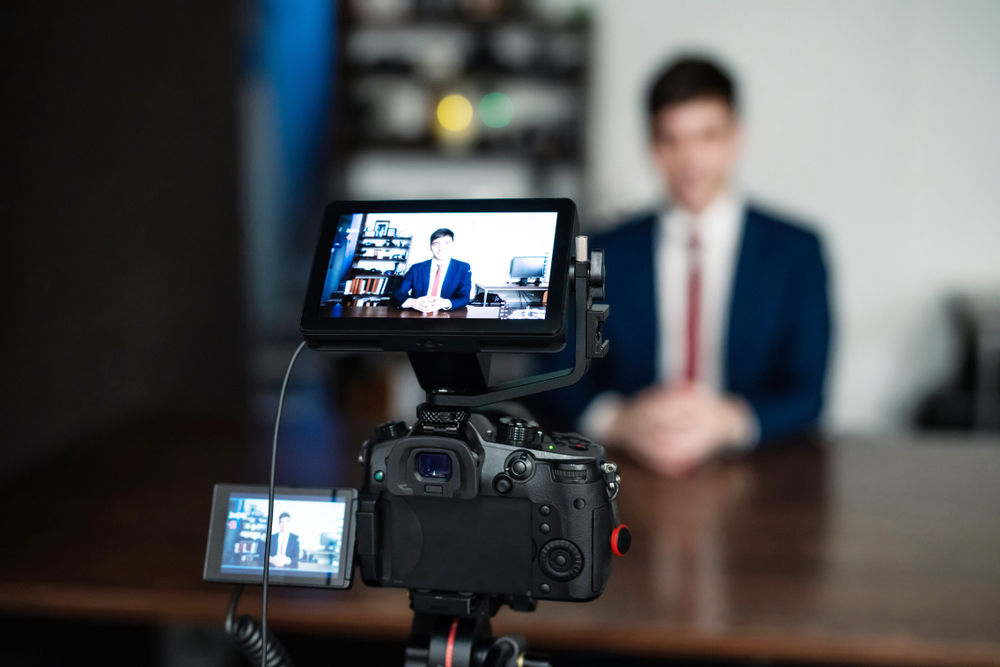 A man in a suit sits before a camera at a desk